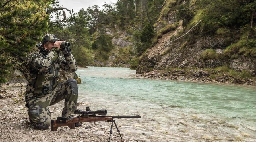 Jäger späht am Ufer mit binokular von Swarovski und Swarovski X5i Zielfernrohr.
