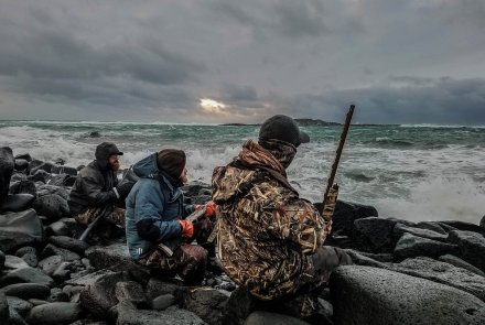 Dream Hunt: Entenjagd in Maine, USA mit Fotograf, Filmemacher und Abenteurer Ryan Baudhuin Ryan Baudhuin und zwei weitere Jaeger kniend auf der Jagd auf Kragenenten auf St. Paul Island, Alaska am Strand vor stuermischer See Rueckansicht