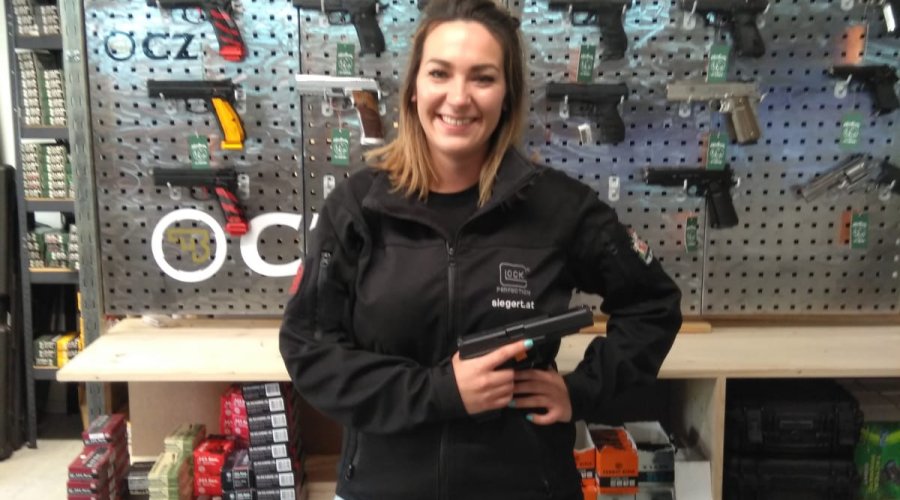 Mitarbeiterin des Shops auf der Schießstätte Zangtal steht vor Regalen mit Kurzwaffen und Munition während sie eine Pistole präsentiert.