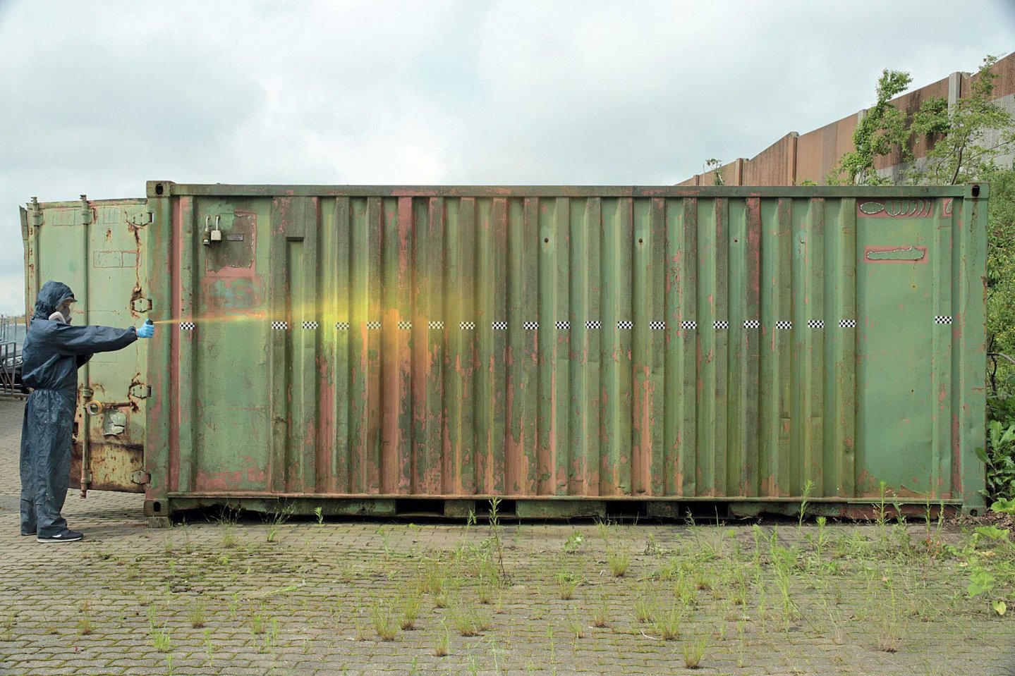 VISIER 8/2016 VISIER-Tester in Schutzkleidung testet Pfefferspray vor einem grünen Container.