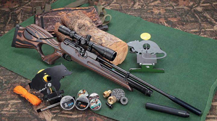 Weihrauch HW 100 T in rechter Seitenansicht an ein Stück Holz gelehnt auf grüner Matte mit Fallscheiben, Munition und Moderator.