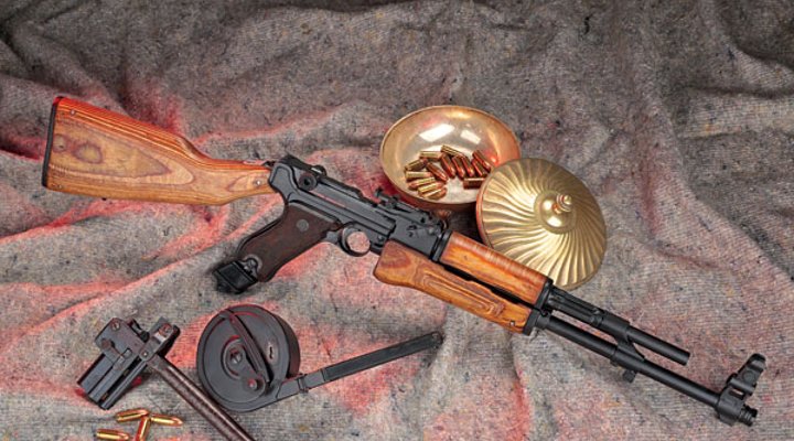 Werle AK-47-Luger in rechter Seitenansicht auf Filztuch mit Trommelmagazin und Patronen in einer Messingschüssel.