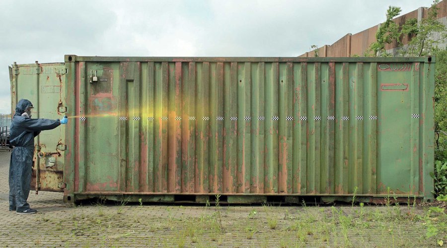 VISIER-Tester in Schutzkleidung testet Pfefferspray vor einem grünen Container.