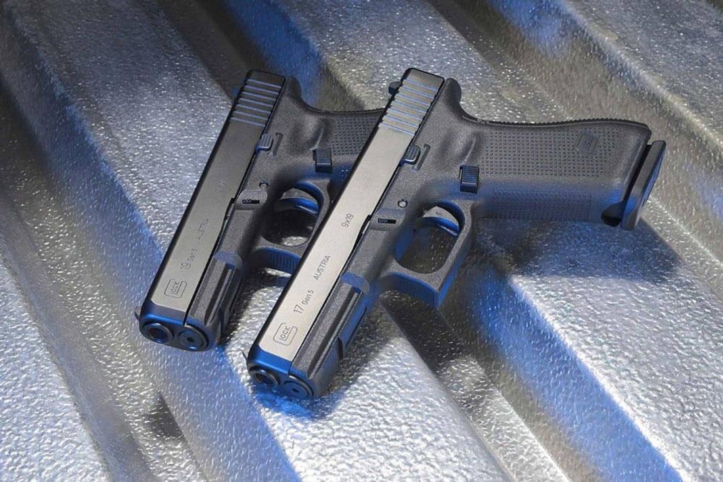 Glock 19 Gen5 (unten) und Glock 17 Gen5 aufeinander in linker Seitenansicht auf Wellblechhintergrund.