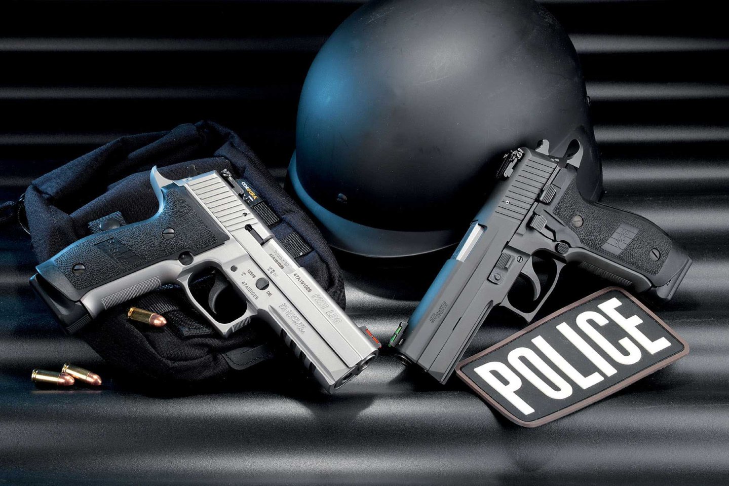 SIG Sauer P 226 LDC II Tacops Stainless in rechter Seitenansicht und SIG Sauer P 226 LDC II Tacops in linker Seitenansicht berühren sich an den Mündungen und bilden ein V. Schwarzer welliger Hintergrund mit schwarzem Helm.