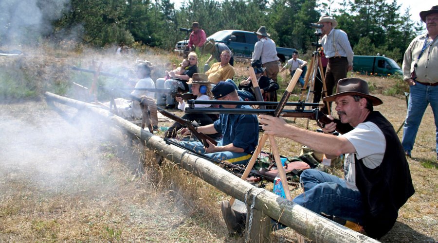 Mehrere Schützen in Westernkleidung schießen ihre historischen Long-Range-Waffen abgestützt auf einen Baumstamm ab.